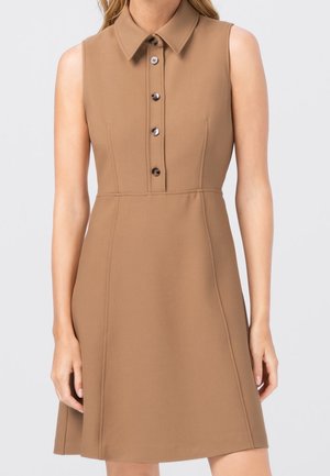 Robe sans manches beige avec col, quatre boutons sur le devant, taille ajustée et jupe trapèze portée par une femme devant un fond uni.