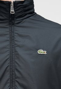 Μπλε σκούρο μπουφάν με φερμουάρ, κατασκευασμένο από λεία ύφη, με ψηλό γιακά και μικρό πράσινο λογότυπο κροκόδειλου στο αριστερό στήθος.