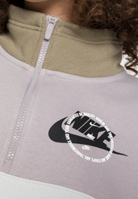 Pullovertrui met een halve rits aan de kraag, lichtpaarse en kaki kleuren, zacht materiaal, en een contrasterend zwart Nike-logo en tekstdetail.