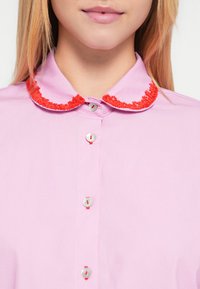 Roosa pluus, millel on punase tikandiga krae, hõbedased nööbid punaste detailidega ja sile tekstuur. Mingeid teisi mustreid ei ole nähtaval.