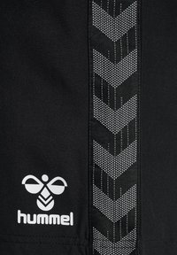 Pantaloni sportivi neri con un motivo a spina di pesce testurizzato su un pannello laterale e logo "hummel" bianco in basso. Tessuto morbido, design atletico.