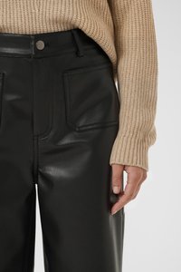Woman wearing black faux leather pants and a beige ribbed knit sweater, hand resting by side, on a plain light background.