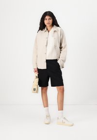 Beige knap-jakke over en cremet kabelstrikket sweater, sorte shorts og beige sneakers. Holder en lille beige taske. Hvid baggrund.