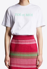 Vit bomulls T-shirt med grön text "TOI et MOI", i kombination med en veckad rosa kjol med flerfärgade horisontella ränder.
