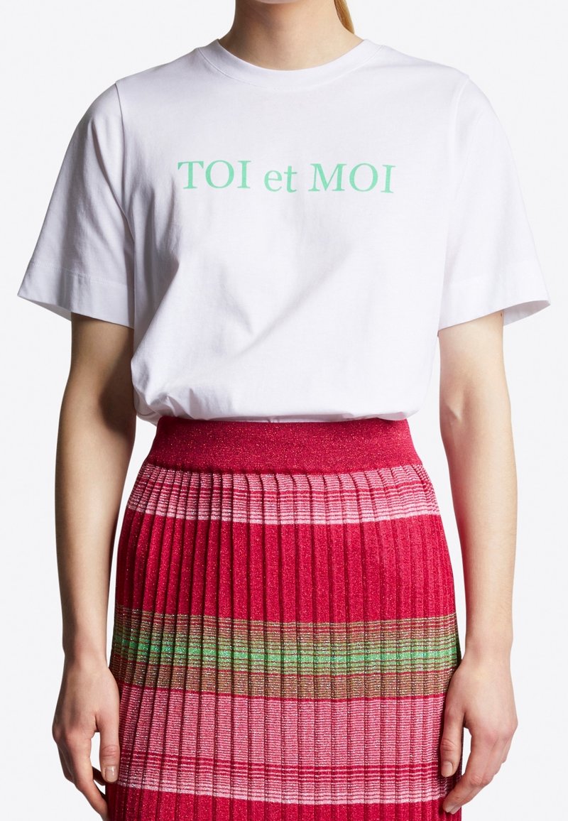 Vit bomulls T-shirt med grön text "TOI et MOI", i kombination med en veckad rosa kjol med flerfärgade horisontella ränder.
