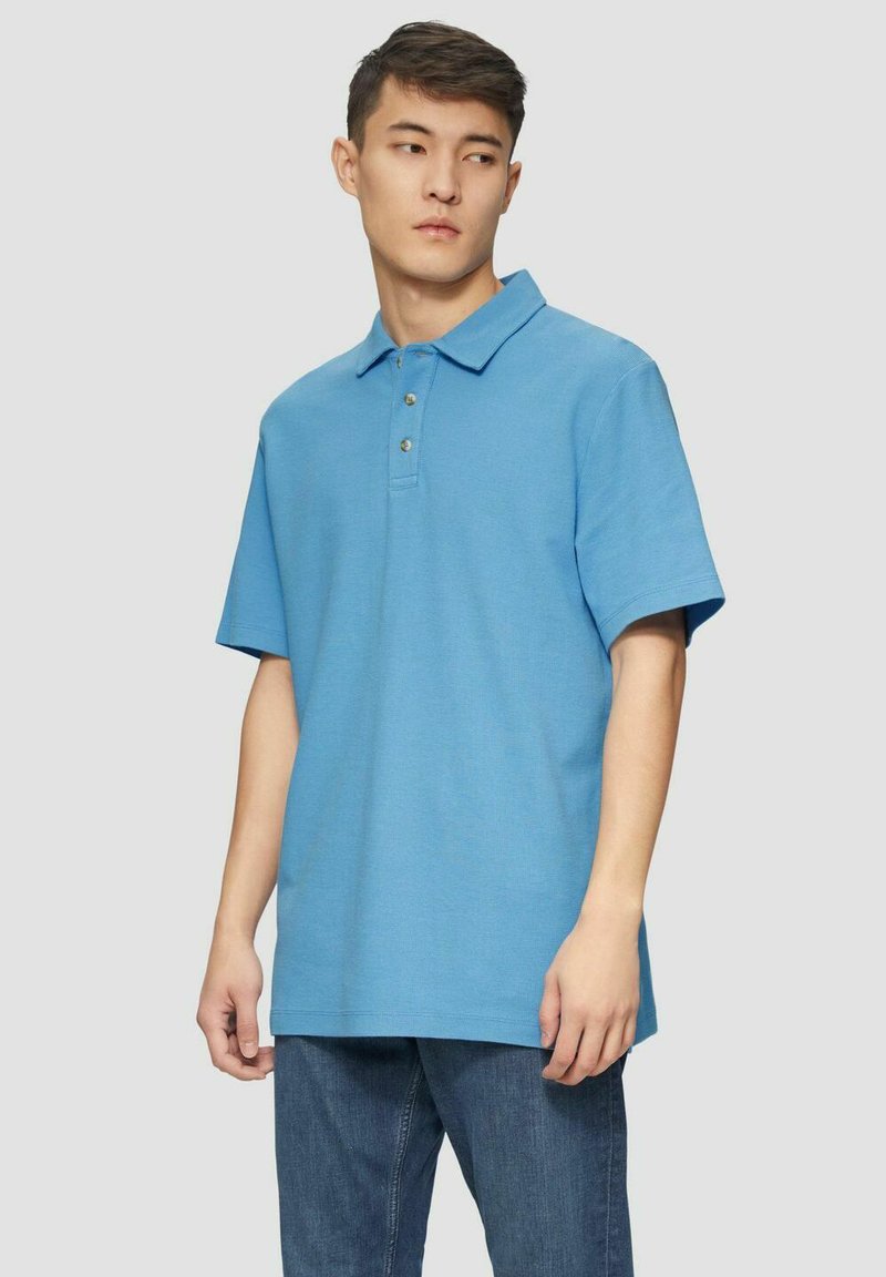 Kurzarm-Poloshirt in Hellblau, aus Baumwolle, mit einem Kragen, drei Knöpfen und einem geraden Saum. Getragen mit einer dunklen Jeans.
