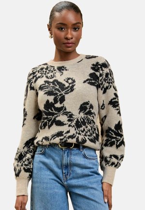 Beige bloemenpull met zwarte patronen, ronde hals en geribbelde manchetten. Gekleed met lichtblauwe high-waisted spijkerbroek en zwarte riem.