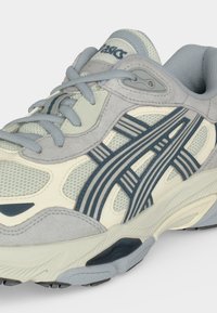 Ανοιχτό γκρι και κρεμ αθλητικό παπούτσι Asics με μπλε λογότυπο, γκρι κορδόνια, επάνω μέρος από δίχτυ και σουέτ και επενδεδυμένη σόλα.