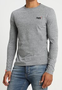 Harmaa, pitkähihaista pehmeästä kankaasta valmistettu t-paita, jossa on pyöreä kaula-aukko, vartalonmyötäinen leikkaus ja pieni logo rinnassa. Yhdistetty sinisiin farkkuihin.