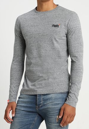 Harmaa, pitkähihaista pehmeästä kankaasta valmistettu t-paita, jossa on pyöreä kaula-aukko, vartalonmyötäinen leikkaus ja pieni logo rinnassa. Yhdistetty sinisiin farkkuihin.