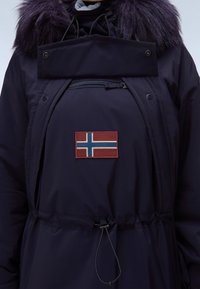 Marinblå vinterjacka med mörk pälskrage, ficka med dragkedja fram, justerbar dragsko i midjan och en röd, blå och vit nordisk flagga patch.