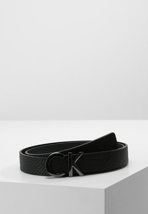 Black textured leather belt coiled on a white surface with glossy black "CK" logo buckle.