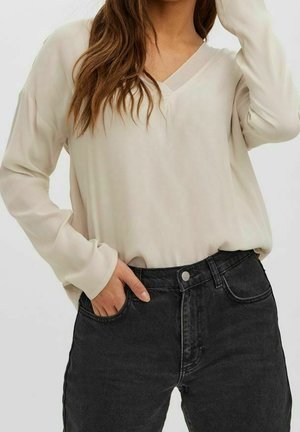 Femme portant un chemisier ample beige clair à manches longues, rentré dans un jean noir taille haute, une main dans la poche et l'autre touchant ses cheveux.