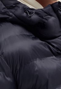 Primer plano de una persona usando una chaqueta de plumas negra con detalles acolchados y un zipper visible en el lado derecho.