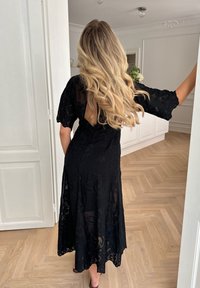 Mustapitsinen mekko kukkakuviolla, lyhyet hihat ja takana avaus. Virtaava siluetti ja nilkkapituinen muotoilu. Taustalla kovapuiset lattiat.