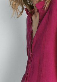 Pink linen button-up shirt with a collar, featuring a relaxed fit, visible buttons, and a slightly open neckline, against a neutral background.