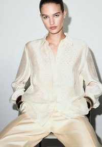 Light beige blouse with button-down collar, subtle polka dot pattern, long sleeves, and a smooth texture, paired with matching satin pants.