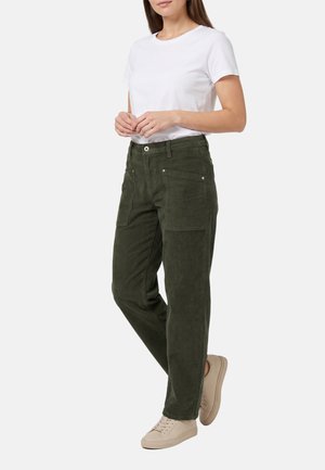 Mujer con una camiseta blanca lisa de manga corta, pantalones verde oliva con grandes bolsillos delanteros y zapatillas beige, de pie con las manos entrelazadas.