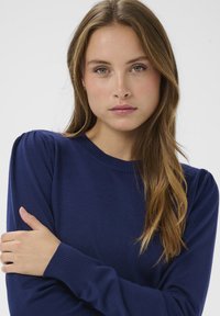 Pull en maille bleu marine avec un col rond, des poignets côtelés et des épaules légèrement bouffantes, présentant une texture douce.