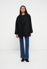 Mulher com cabelo encaracolado, vestindo um casaco oversized preto, um suéter escuro, jeans azuis e mocassins pretos, de pé contra um fundo branco simples.