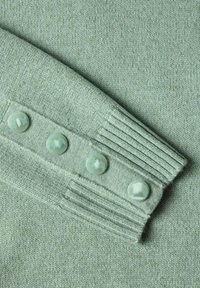 Gros plan d'une manche de pull tricoté vert clair avec quatre boutons ronds assortis le long du poignet avec détails côtelés.