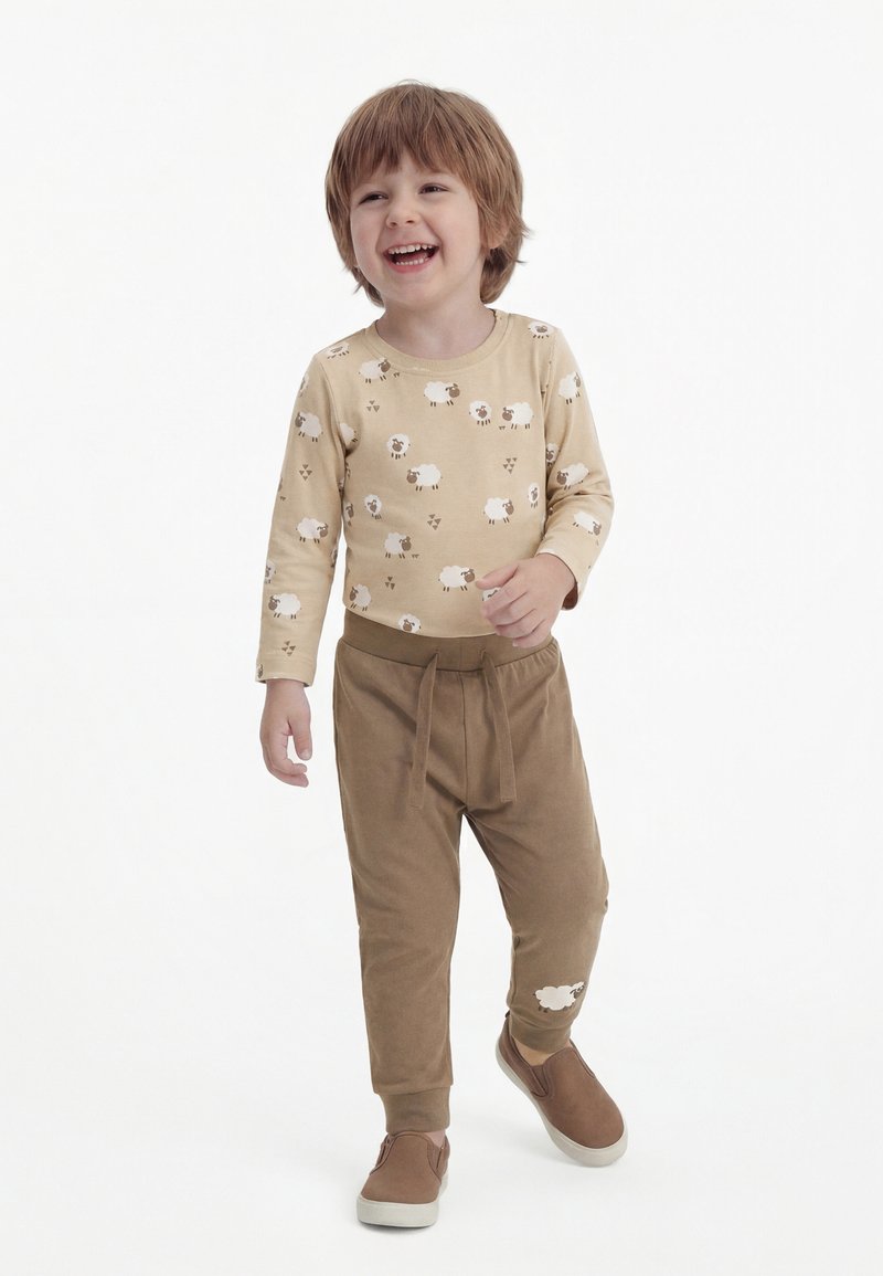 Jeune enfant souriant, portant une chemise beige à manches longues avec des motifs de moutons, un pantalon marron avec un détail mouton, et des chaussures marron à enfiler, debout sur un fond blanc.