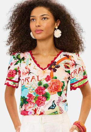 Kvinde med krøllet hår iført store hvide blomsterøreringe og en floral V-hals-top med røde kanter samt hvide bukser.