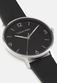 Montre analogique noire avec un boîtier en acier inoxydable argenté, un bracelet en caoutchouc et un cadran minimaliste comportant des chiffres et des marqueurs de minutes, avec le nom de la marque visible.