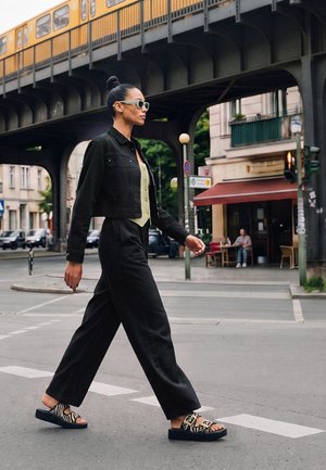 Femme traversant la rue sous des voies ferrées jaunes surélevées en milieu urbain, portant une tenue noire et des sandales à motifs.