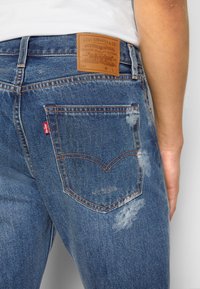 Gros plan de la poche arrière bleue d'un jean Levi's avec des coutures visibles, des taches décolorées et un patch en cuir marron sur la ceinture.