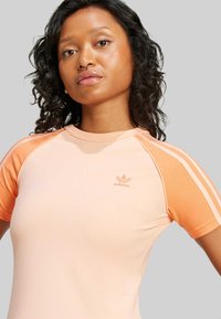 Persikø og orange kortærmet træningsshirt med kontrasterende ærmer, glat tekstur og broderet Adidas-logo på brystet.