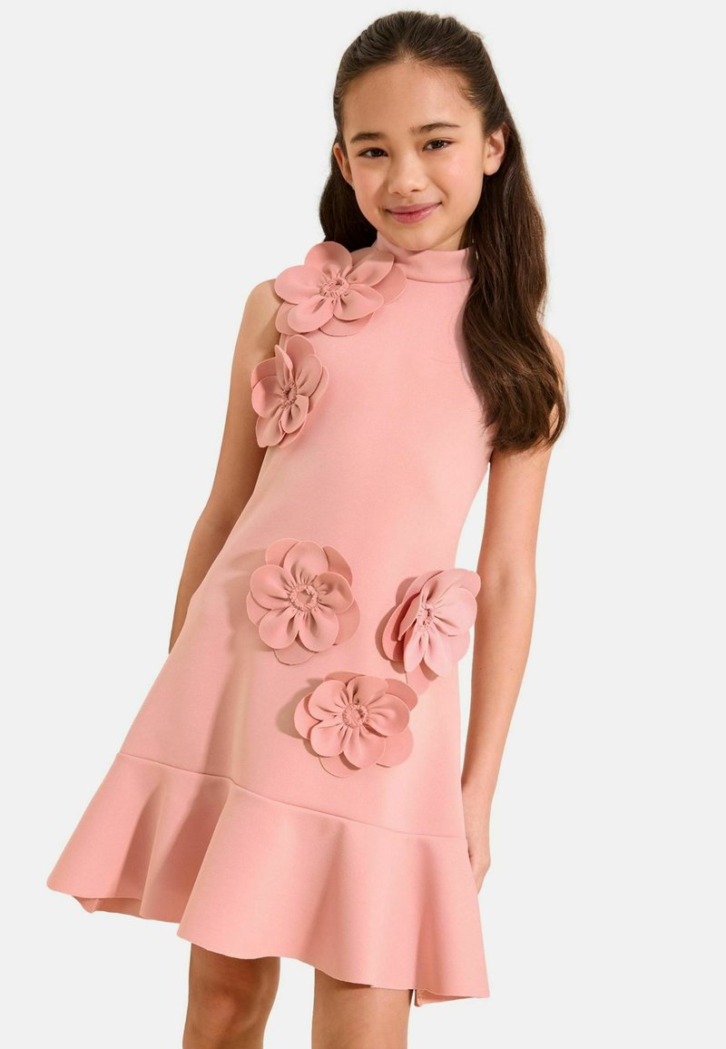 Mädchen mit langen braunen Haaren, das ein ärmelloses pinkfarbenes Kleid mit großen 3D-Blumenapplikationen und einem ausgestellten Saum trägt, leicht lächelnd.