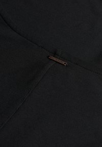 Nahaufnahme einer schwarzen Stoffnaht mit einem kleinen rechteckigen Metallanhänger in der Mitte, auf dem "Superdry" steht.