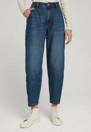Frau trägt blaue Jeans mit hoher Taille und zuspitzenden Beinen, ein beiges langärmliges Oberteil und weiße hohe Sneakers, steht mit einer Hand in der Tasche.