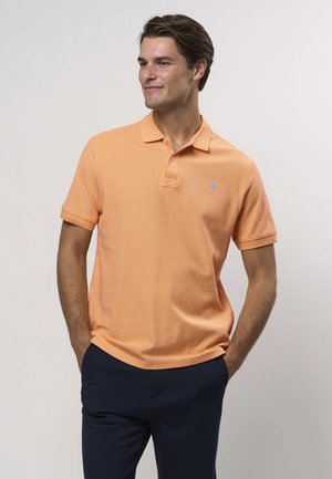 Tricou polo portocaliu cu guler clasic, mâneci scurte, material texturat și un mic logo albastru pe pieptul stâng. Asortat cu pantaloni închiși la culoare.