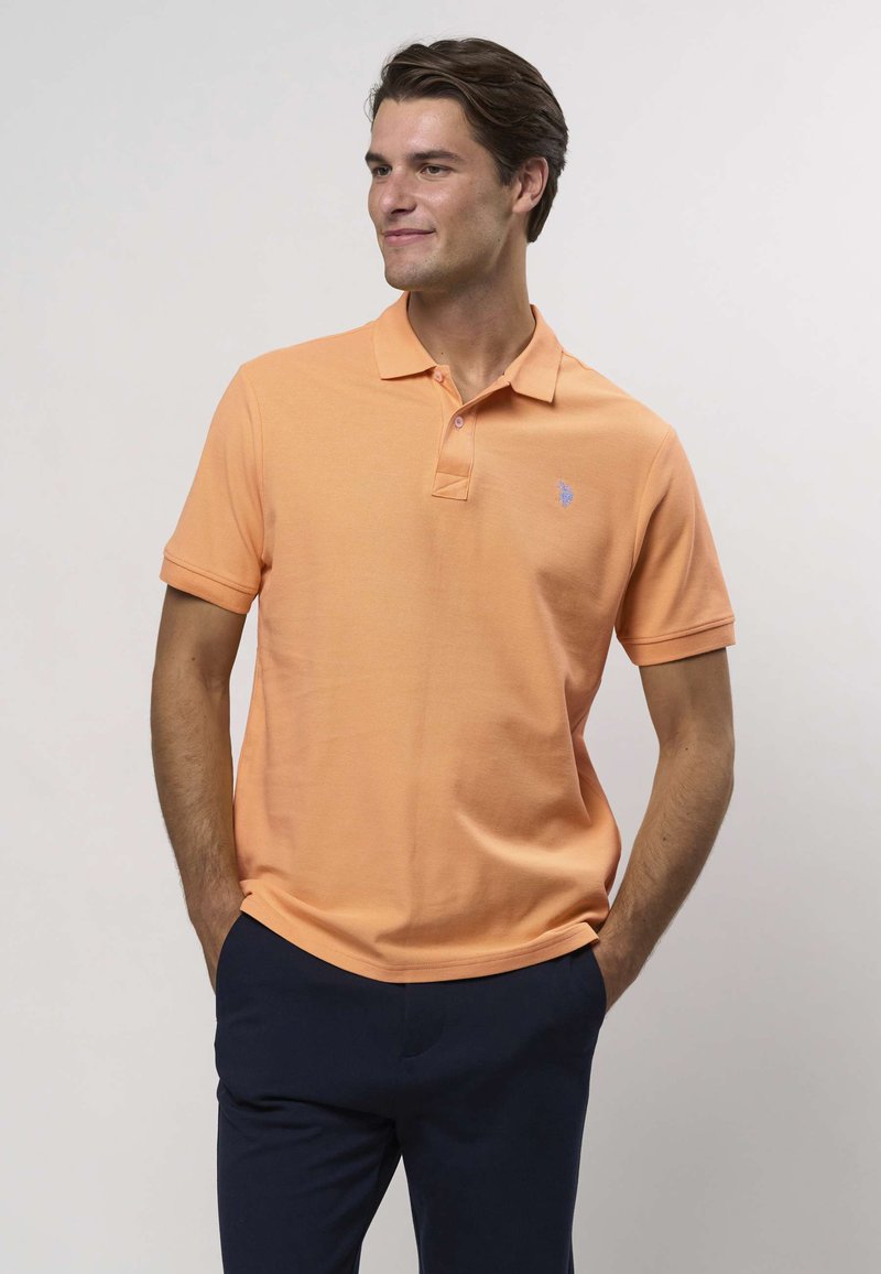 Oranž polo-särk klassikalise kraega, lühikeste varrukatega, tekstuurse kangaga ja väikese sinise logoga vasakul rinnal. Ühildatud tumedate pükstega.