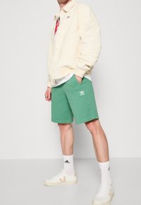 Světle krémová bunda, zelené kraťasy s logem, bílé tenisky s béžovými akcenty a bílé ponožky. Neformální outfit na jednolitém pozadí.