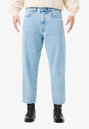 Uomo che indossa jeans azzurri a gamba dritta con tasche frontali e stivali neri con punta quadrata, in piedi con le braccia leggermente distanziate dai fianchi.