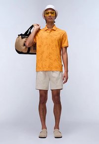 Polo arancione con motivo a foglie, pantaloncini beige e scarpe slip-on di colore chiaro. In mano una borsa da viaggio nera e marrone chiaro. Cappello bianco e occhiali da sole gialli.