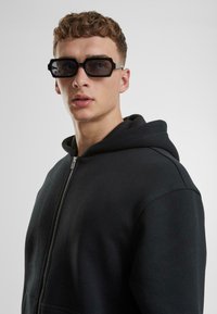 Haut à fermeture éclair noir en tissu doux, avec une capuche spacieuse. Le modèle porte des lunettes de soleil noires rectangulaires au design angulaire.