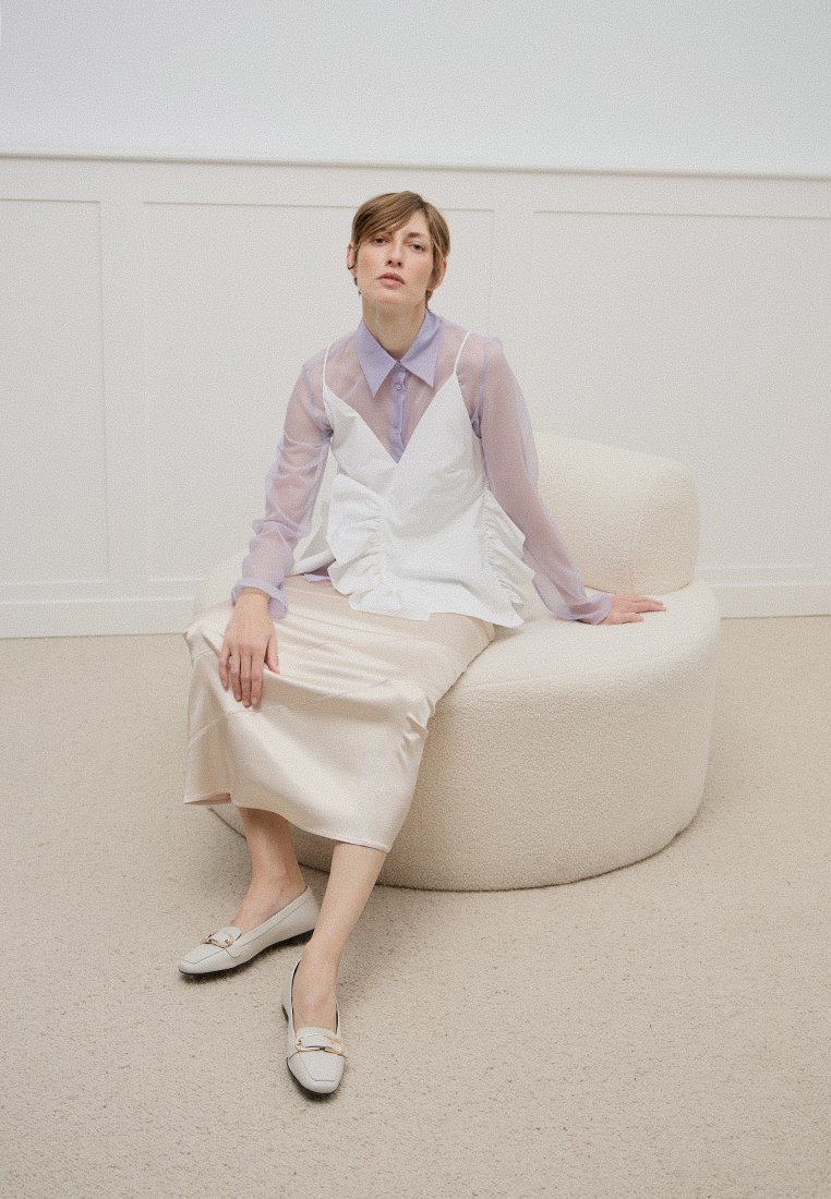Weißes ärmelloses Oberteil mit Rüschen über einer helllila durchsichtigen Bluse; cremefarbene Satin-Rock und graue Loafers. Sitzt auf einem runden, weichen Stuhl.