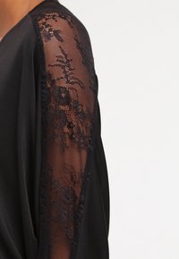 Svart plagg med genomskinliga spetsärmar med blommönster, en mjuk textur och en lös passform längs kroppen.