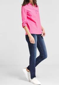 Camicia button-up rosa brillante con maniche lunghe, abbinata a jeans skinny blu scuro e sneakers bianche. Presenta un dettaglio della tasca frontale.