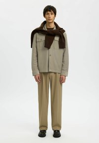 Man in beige jack en broek, met een donkerbruine trui over zijn schouders en zwarte schoenen, staande tegen een witte achtergrond.