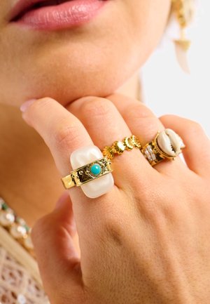 Bagues en or et argent avec différents motifs : l'une présente une pierre givrée et de la turquoise, une autre des accents festonnés, et une avec un motif de coquillage.