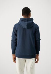 Námořnická modrá mikina s kapucí na šňůrku, s žebrovanými manžetami a spodním lemem, vyrobená z hladké bavlněné látky. Nosená s světlými kalhotami.