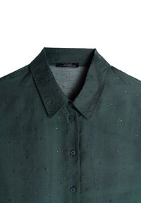Ciemnozielona koszula o miękkiej teksturze, z guzikiem z przodu i małymi, błyszczącymi zielonymi detalami rozsianymi po tkaninie.