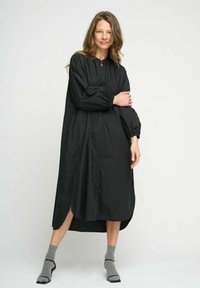 Rochie neagră, cu mânecă lungă, cu nasturi în față și fente laterale, dintr-un material ușor. Purtați cu șosete gri și sandale minimaliste.