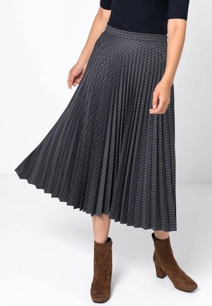 Vrouw draagt een donkere geplooide midi-rok met een geruit patroon, zwarte top en bruine suède enkellaarsjes, staand op een witte achtergrond.
