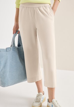 Kvinde iført beige cropped bukser, hvide sneakers med blå accenter, holder en stor lyseblå denim tote-taske, hånd i lommen.